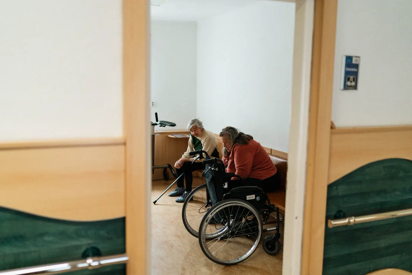 Two older women sit together in a nursing home lounge, one using a wheelchair.