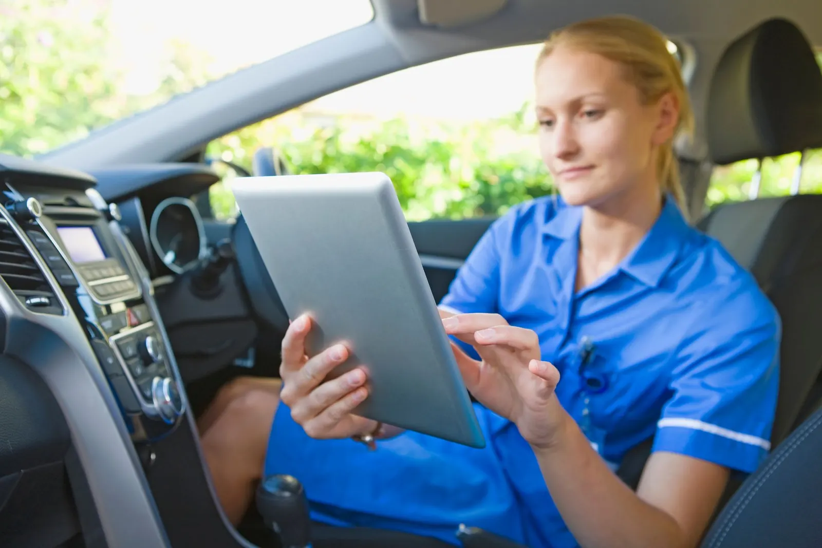Un membre du personnel infirmier est assis au volant d’une voiture et utilise une tablette.